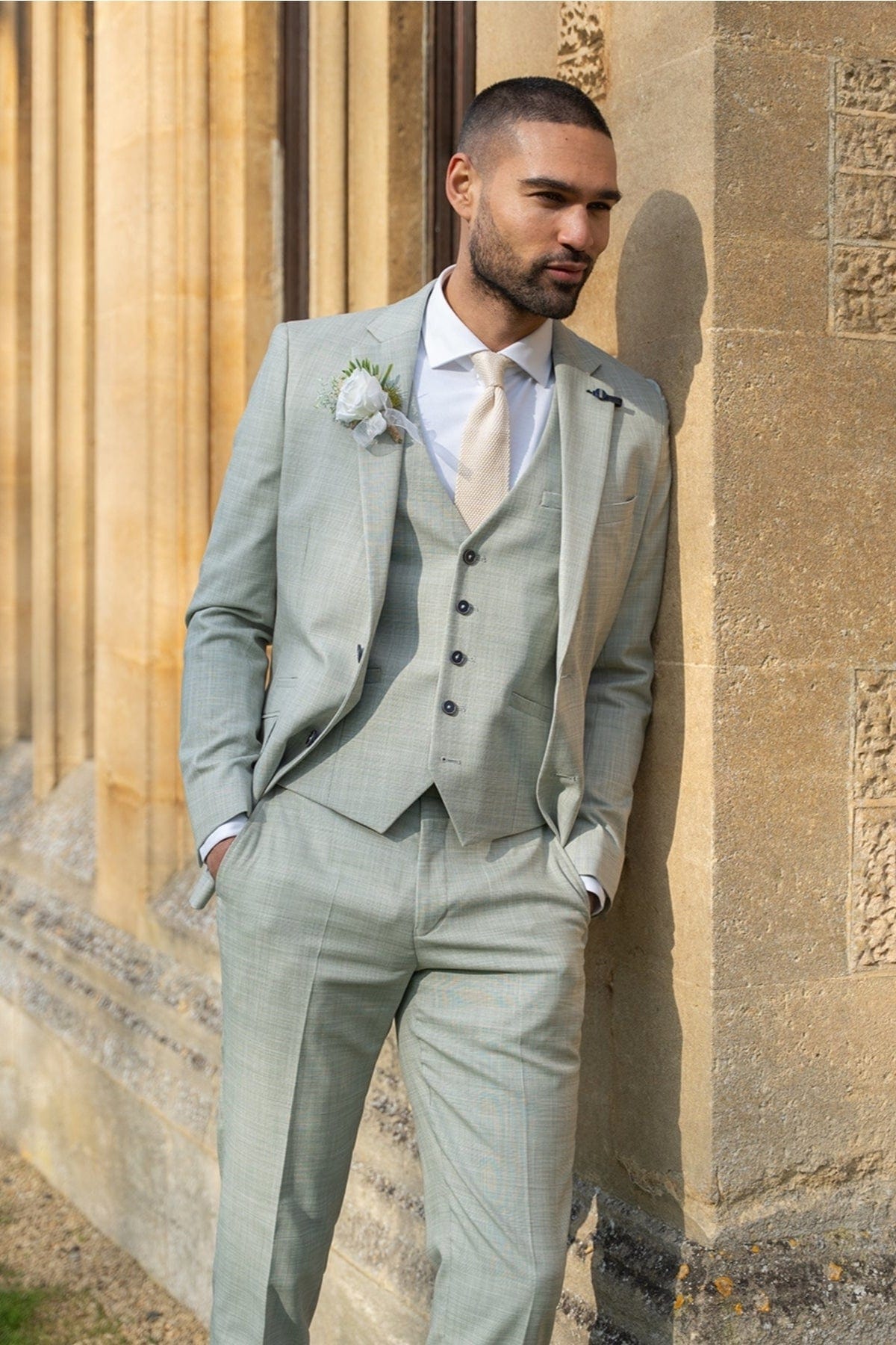 Man in a light sage green suit standing against a stone wall.
