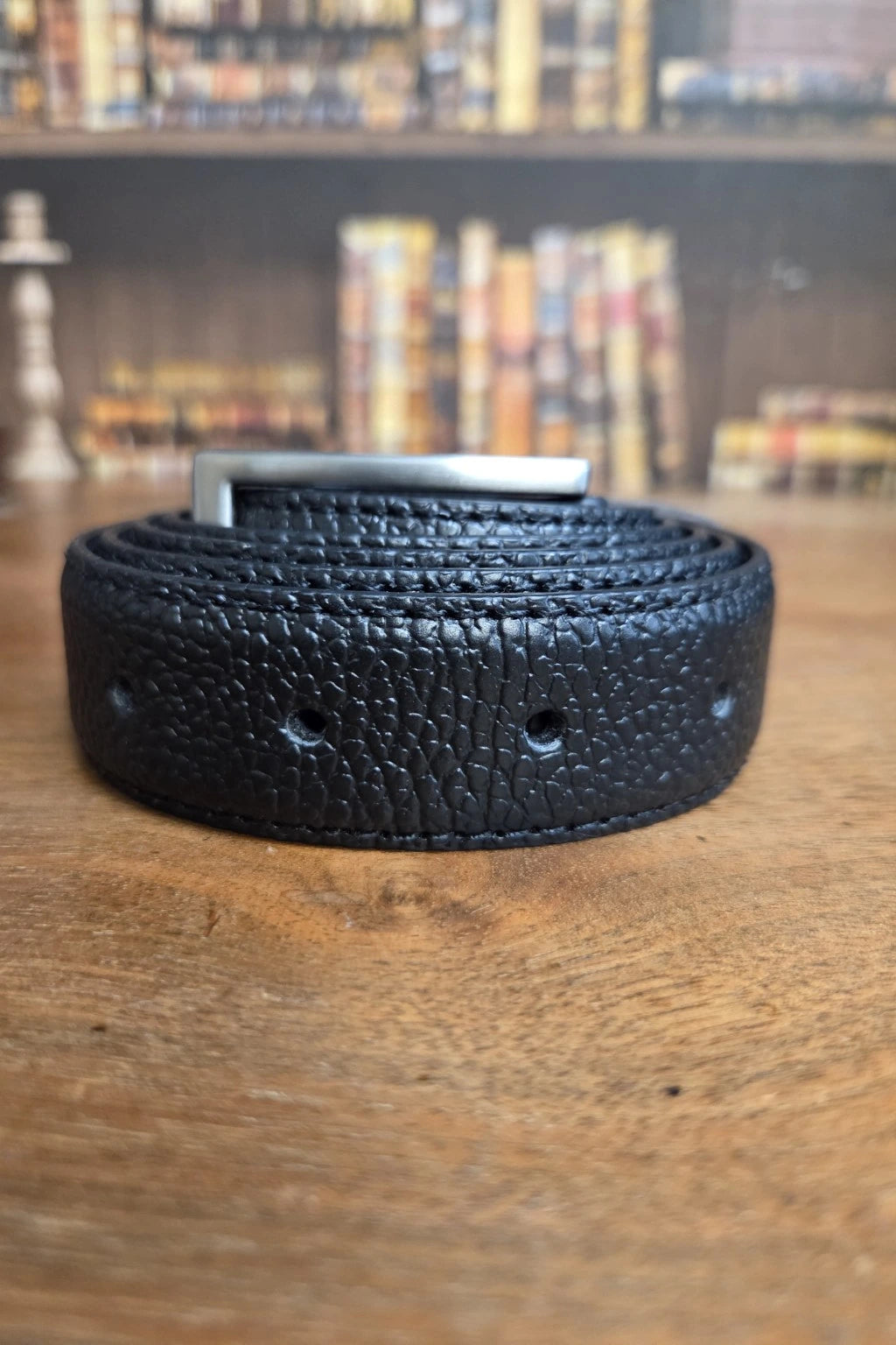 A black textured leather belt with a silver metal buckle, coiled on a wooden surface with a blurred bookshelf background.