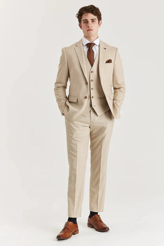 A male model wearing a beige three-piece suit with a waistcoat and trousers, paired with a white shirt and brown shoes.