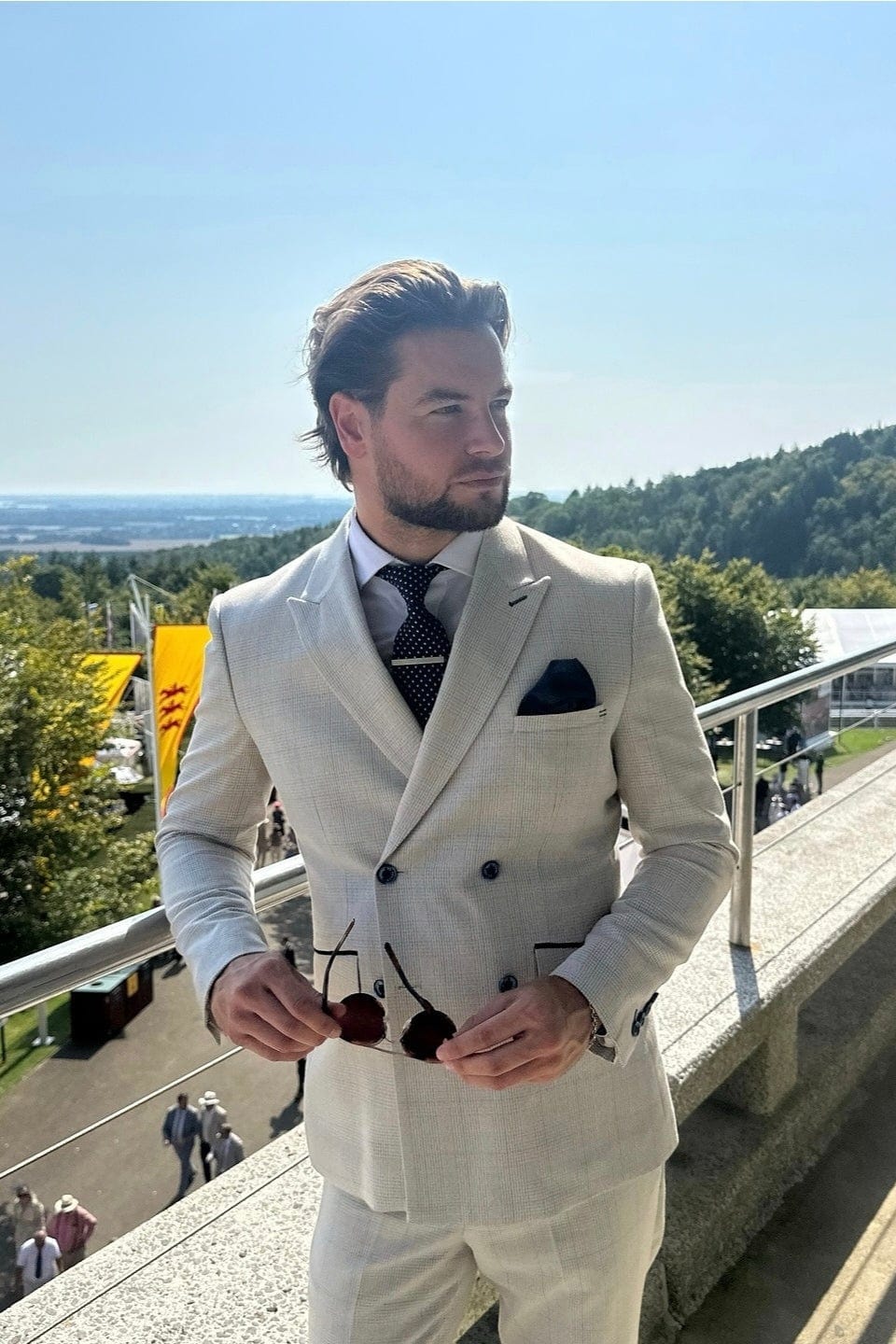 Man in a light-colored suit standing on a balcony with a scenic view.