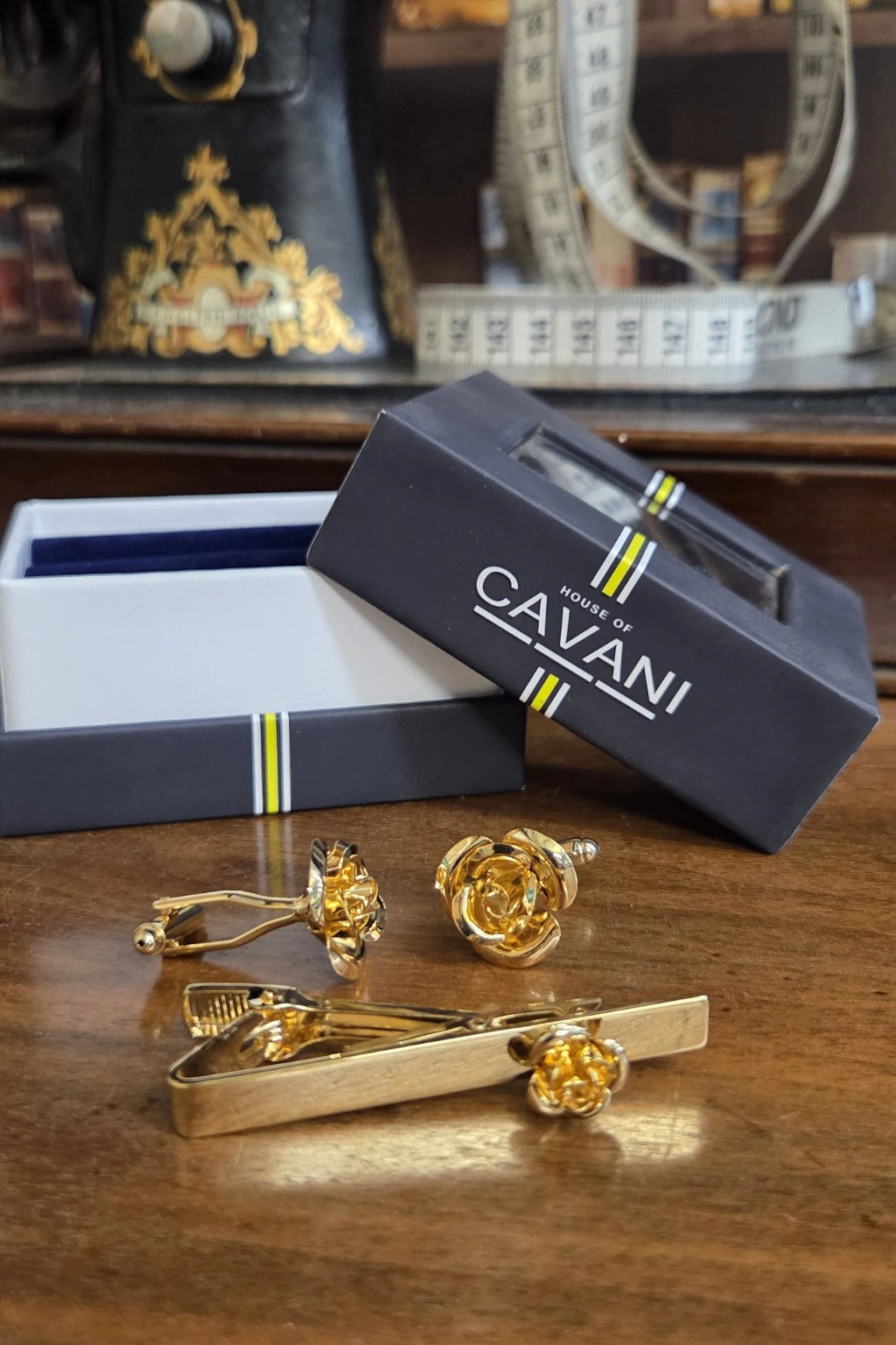 A gold rose-themed men's accessory set including a tie clip, tie clip bar, and cufflinks, displayed on a wooden table next to a dark gray CAVANI brand gift box.