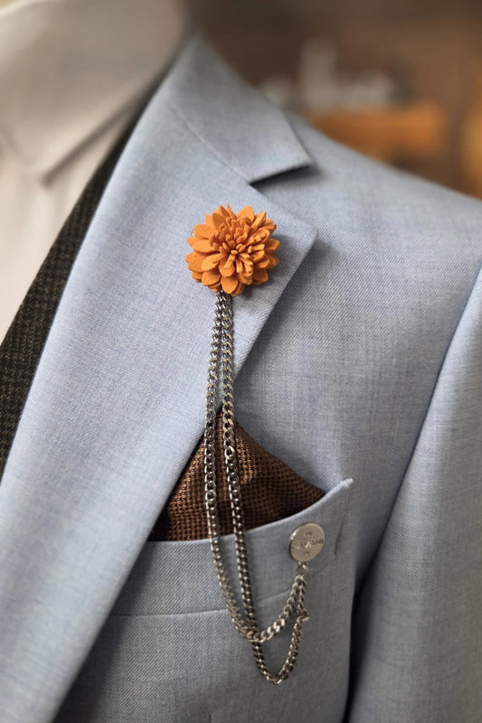 An orange fabric flower lapel pin with a silver chain and ball chain accessory, pinned to a light blue suit jacket over a brown patterned pocket square.