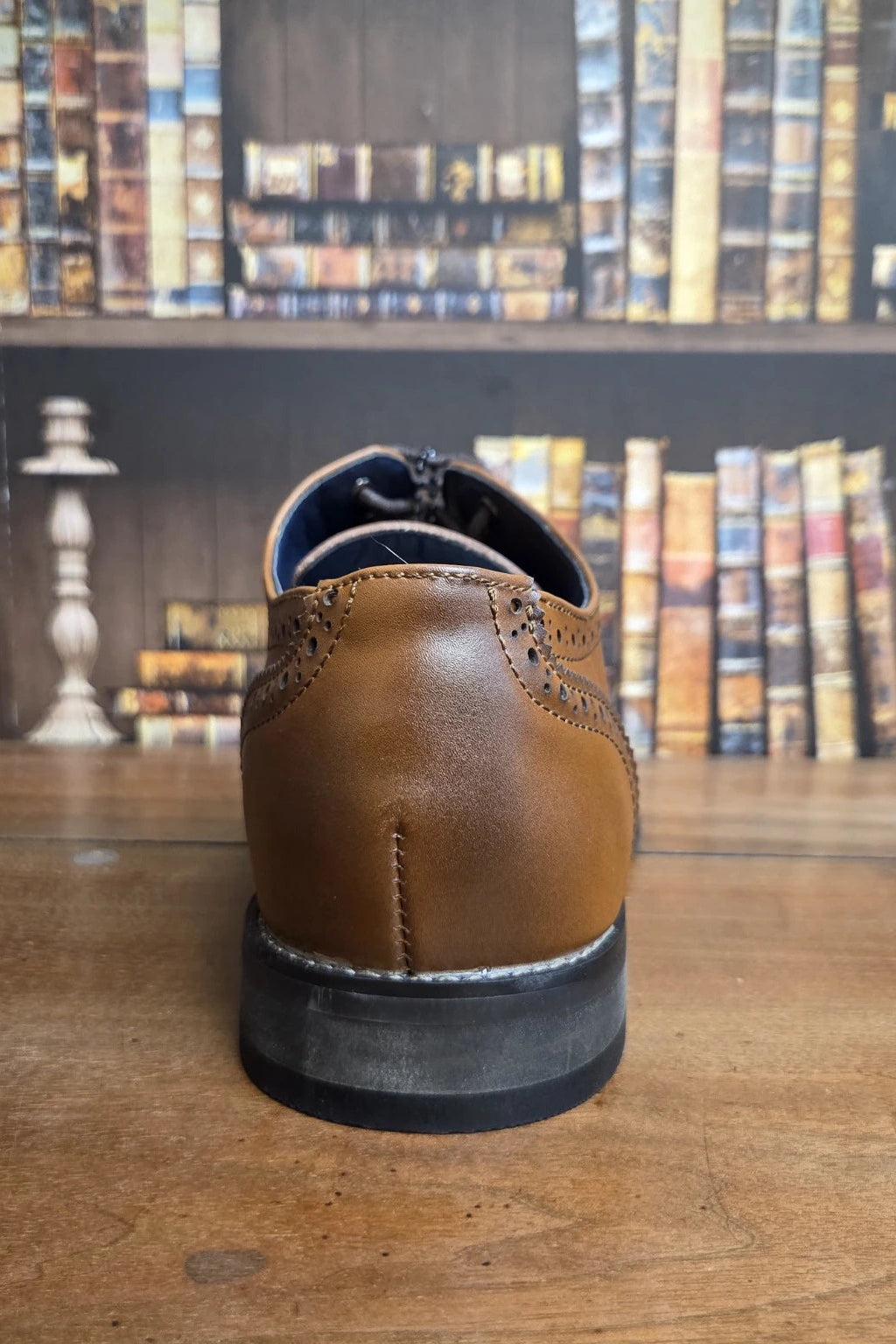 A brown leather wingtip shoe shown from the back, positioned on a wooden table with a blurred bookshelf background.