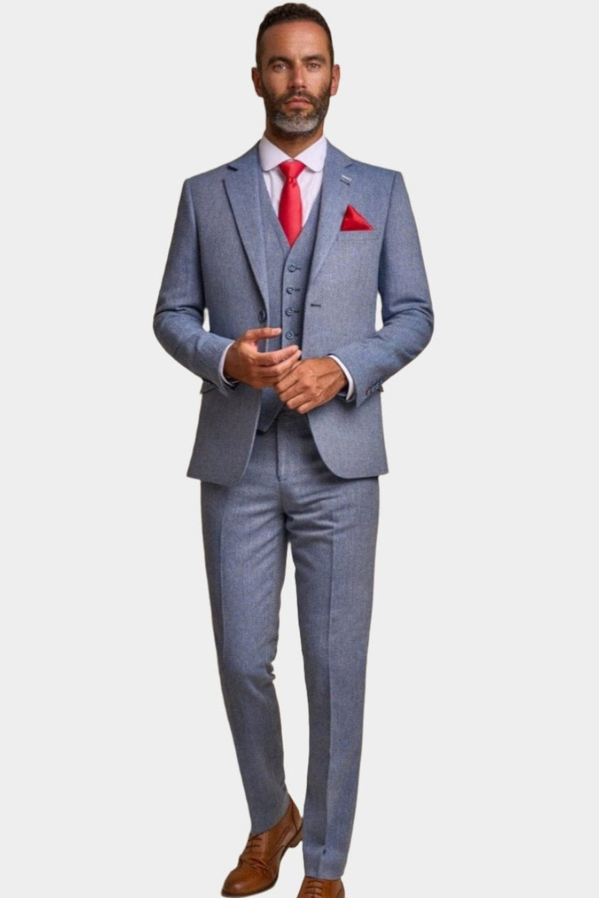 A man wearing a blue tweed three-piece suit with a red tie and a pocket square, standing against a white background.
