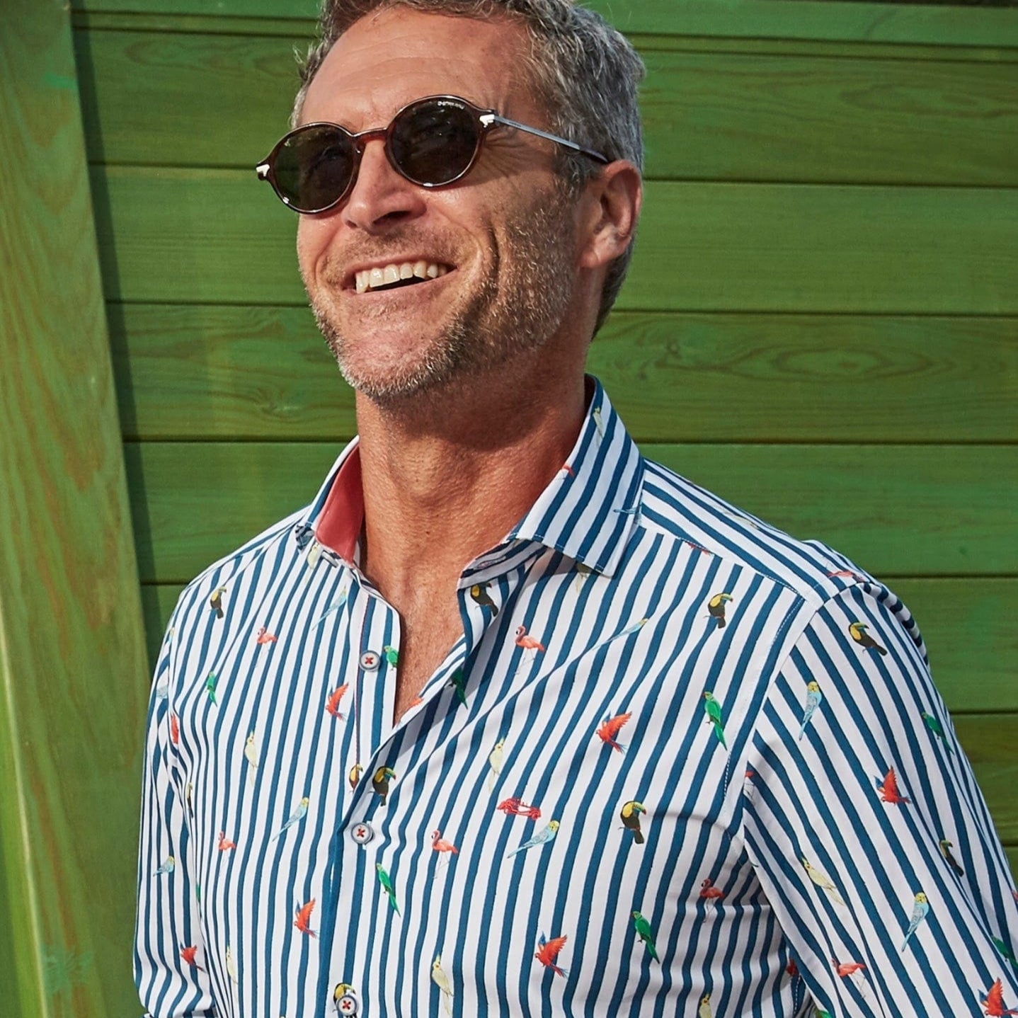 A man wearing a long-sleeved, striped navy and white Dario Beltran shirt with a tropical bird pattern, standing in front of a green wall.