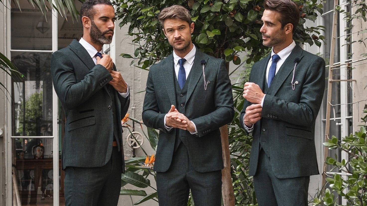 Three men in Cavani Caridi Olive three-piece suits standing outdoors with plants in the background