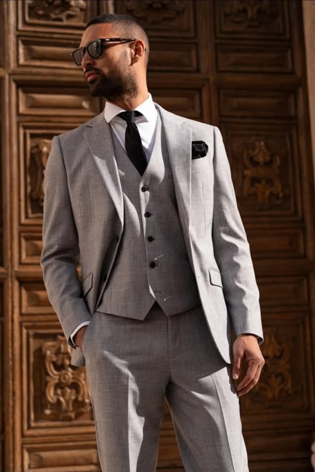 A man wearing the Cavani Malibu grey three-piece suit, white dress shirt, black tie, and black sunglasses, standing in front of a dark brown carved wooden door.
