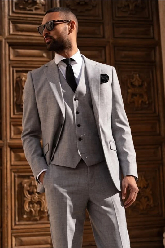 A man wearing the Cavani Malibu grey three-piece suit, white dress shirt, black tie, and black sunglasses, standing in front of a dark brown carved wooden door.