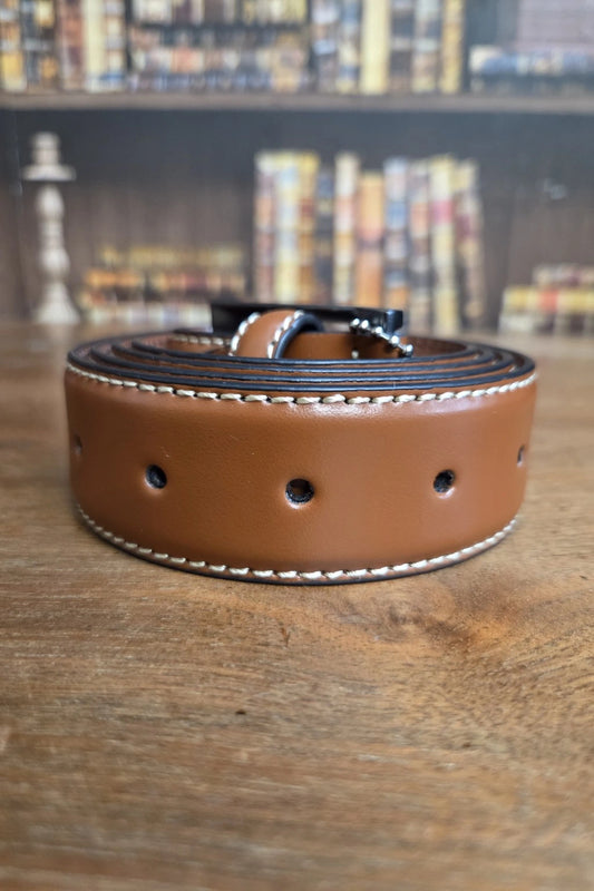 A rolled brown leather belt with white contrast stitching and a silver buckle, resting on a wooden tabletop with a blurred bookshelf background.