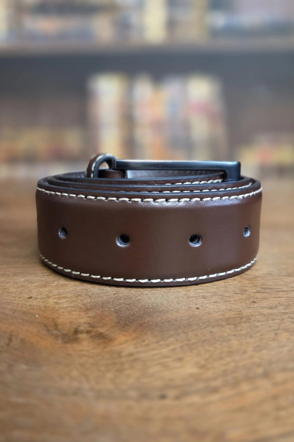 A dark brown leather belt with white contrast stitching and a black metal buckle, coiled on a rustic wooden surface with a blurred background.