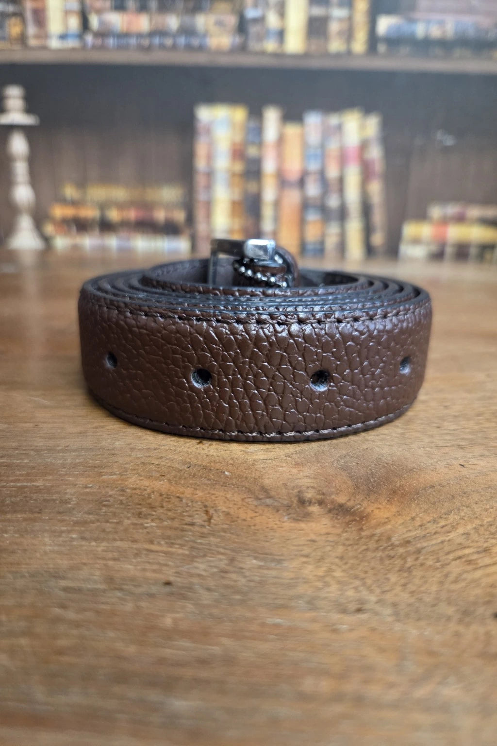 A dark brown textured leather belt with a silver buckle, coiled on a wooden tabletop with a blurred bookshelf background.