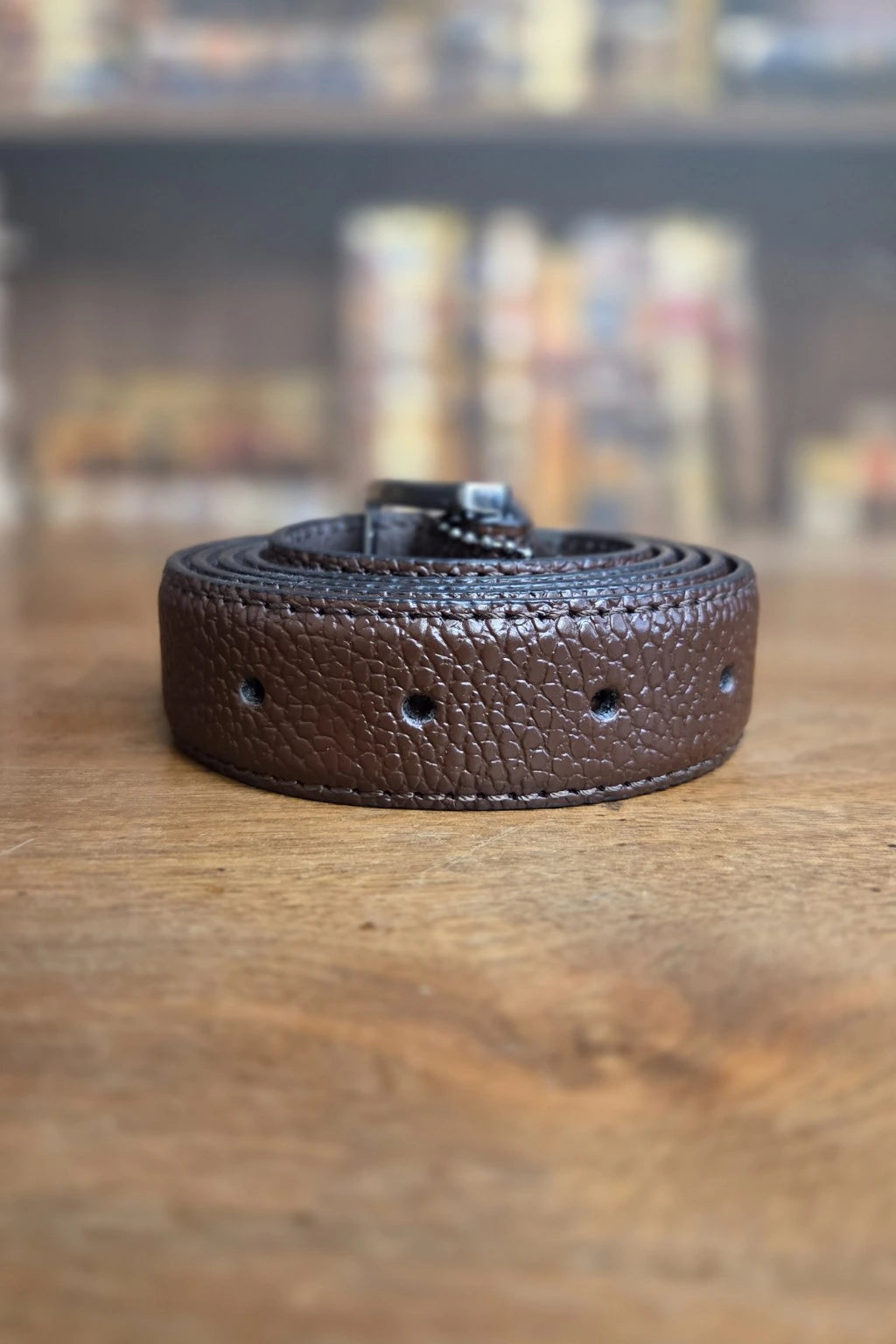 A dark brown textured leather belt with a black buckle, coiled on a wooden surface with a blurred background.