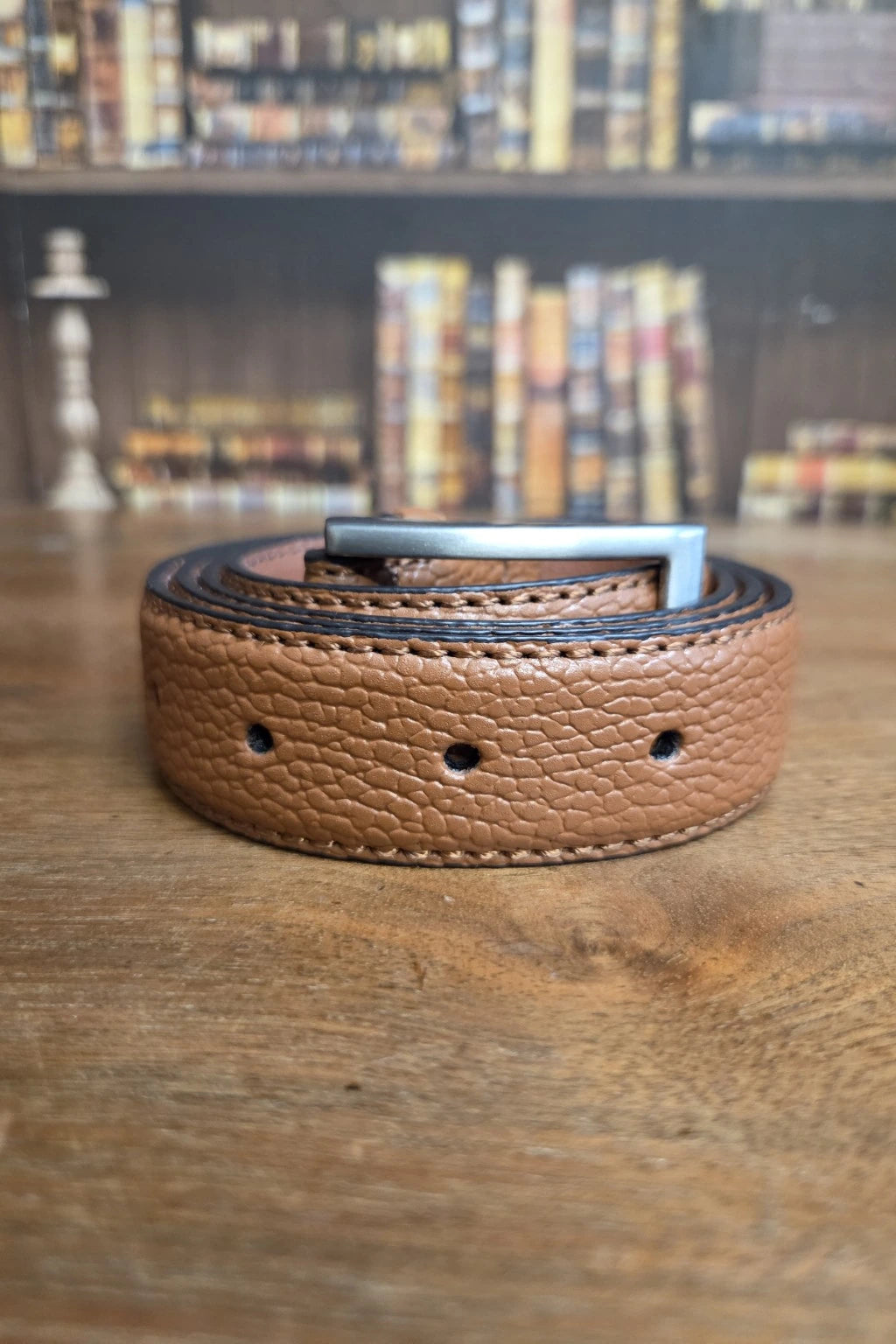 A tan pebbled leather belt with a silver metal buckle, coiled on a wooden table with a blurred bookshelf background.