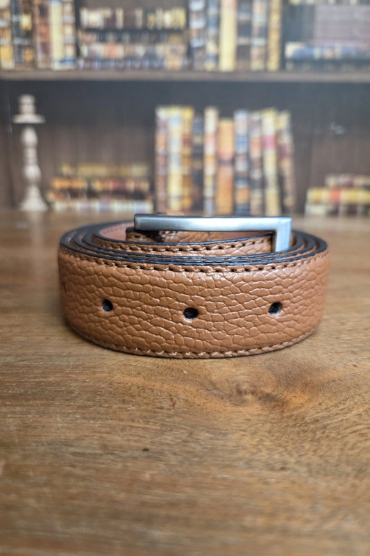 A tan pebbled leather belt with a silver metal buckle, coiled on a wooden table with a blurred bookshelf background.