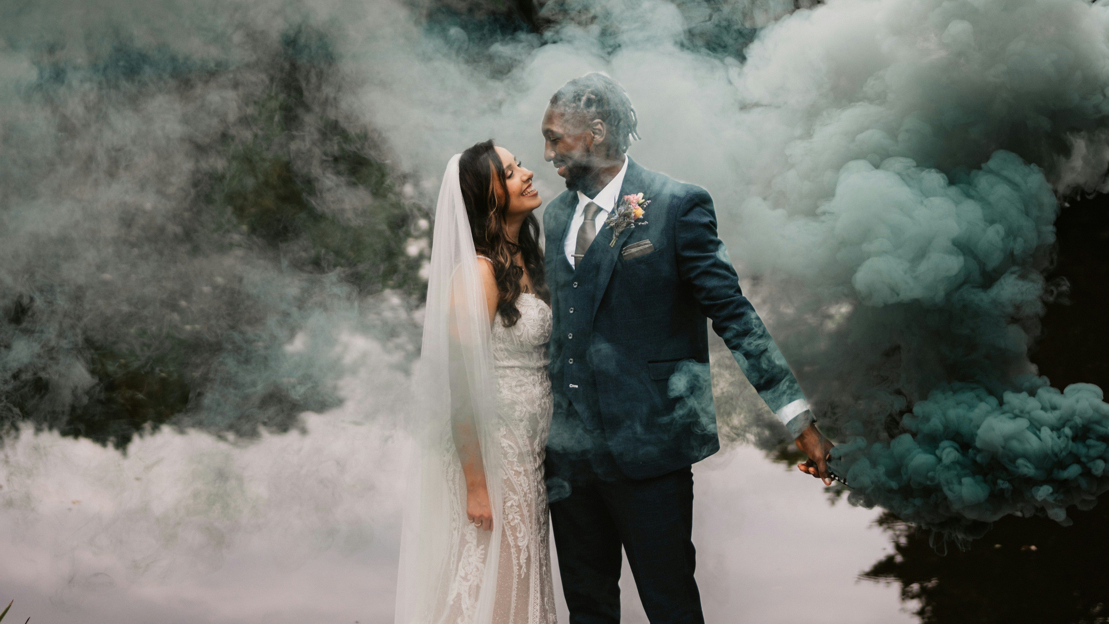 Bride and groom, celebrating there wedding day, smiling at each other with smoke and green flares. the groom in wearing a caridi olive 3 piece suit on his wedding day.