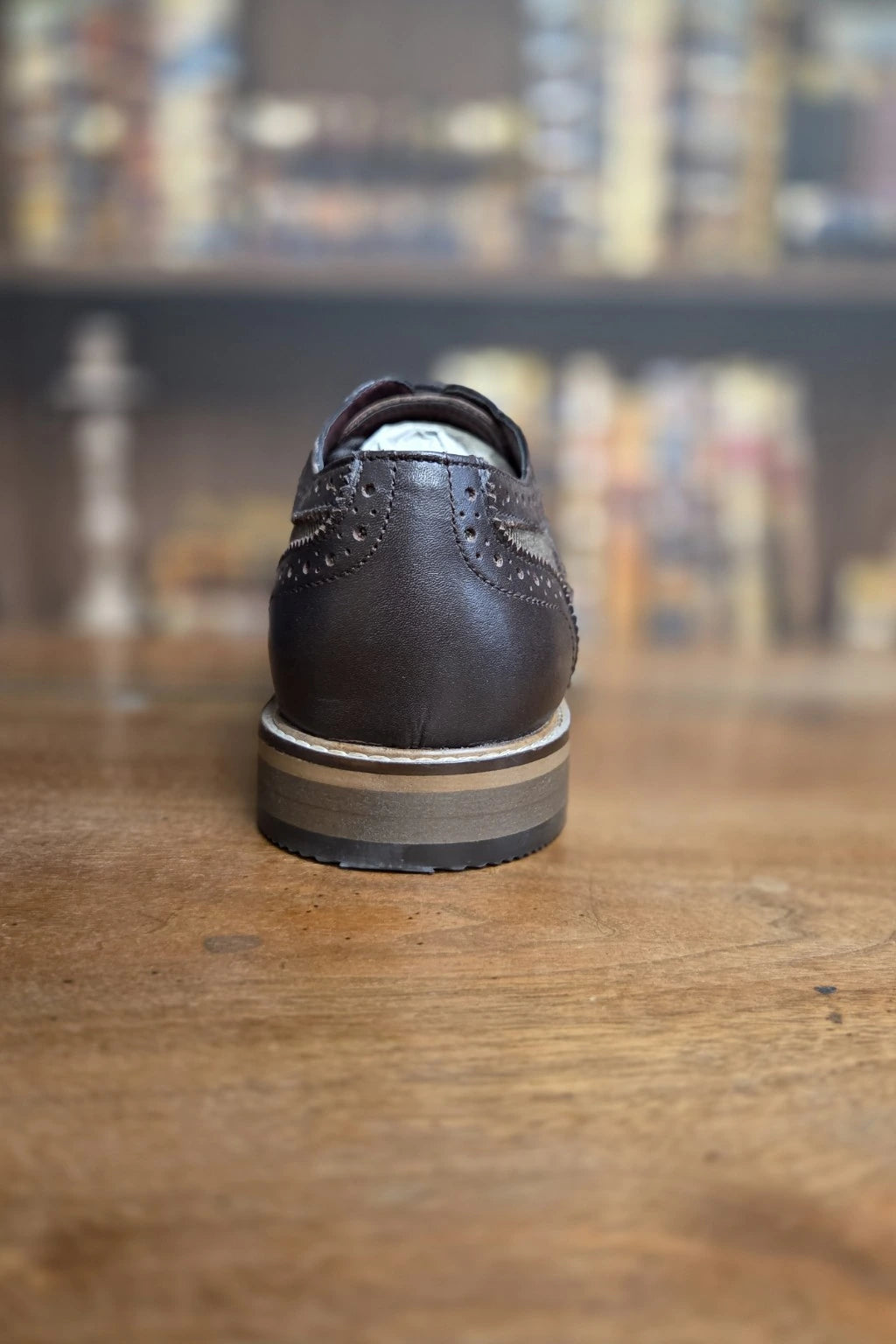 A Cavani Ellington leather wingtip shoe shown from the back, resting on a wooden table with a blurred bookshelf background.