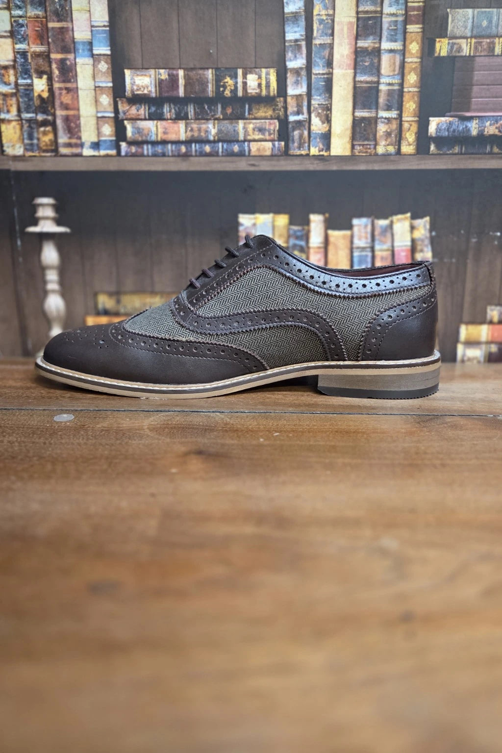 A Cavani Ellington brown brogue wingtip shoe with a textured patterned upper, resting on a wooden table with a bookshelf in the background.