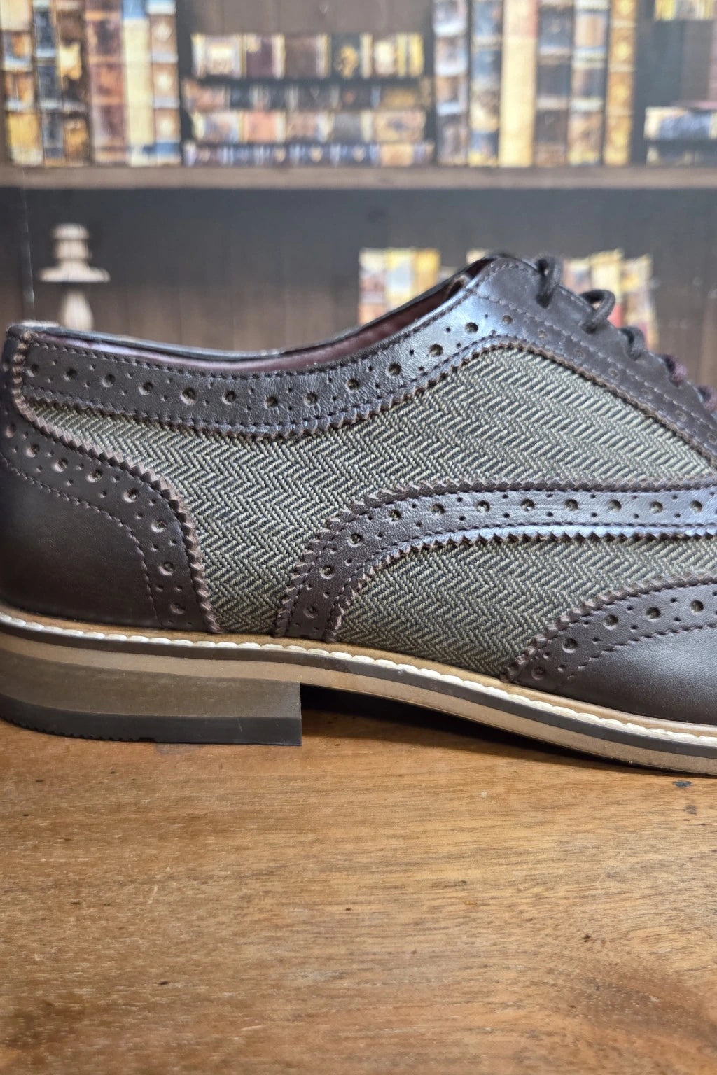 A Cavani Ellington brown leather and herringbone-patterned wingtip brogue shoe resting on a wooden surface with a blurred bookshelf background.