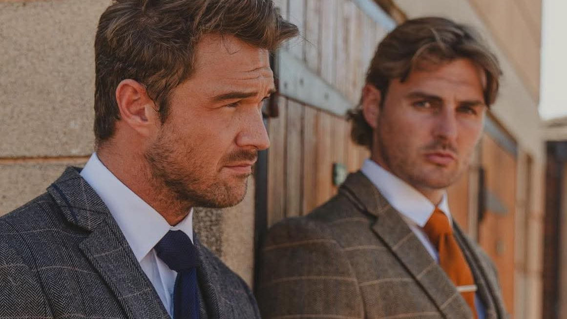 Two men in House Of Cavani race day suits standing side by side against a building.