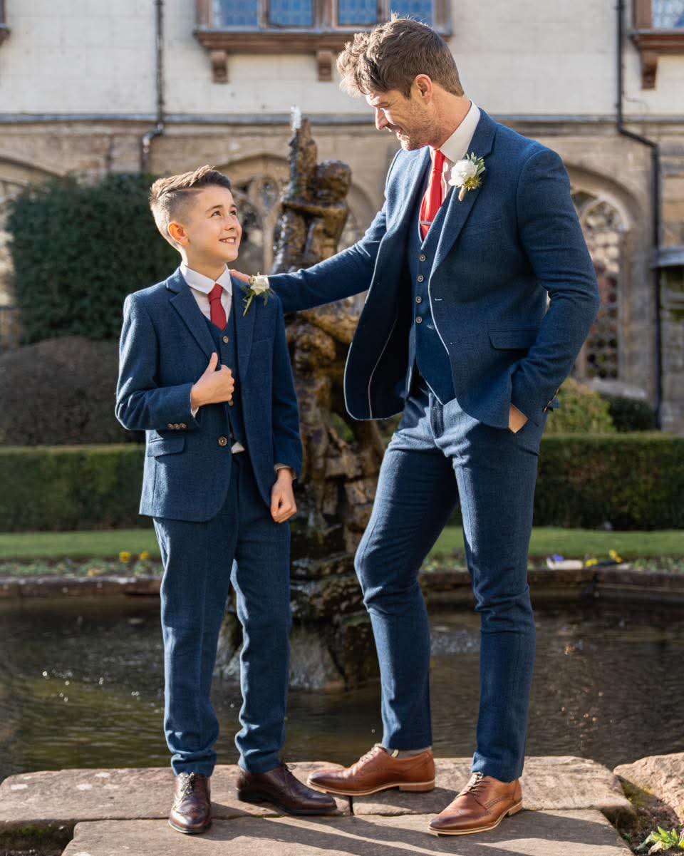 A father and his son in matching blue tweed suits standing outdoors at a wedding with a fountain in the background.