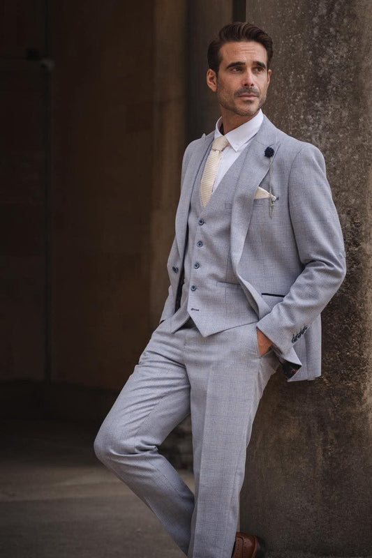Man in a light blue suit leaning against a textured wall.