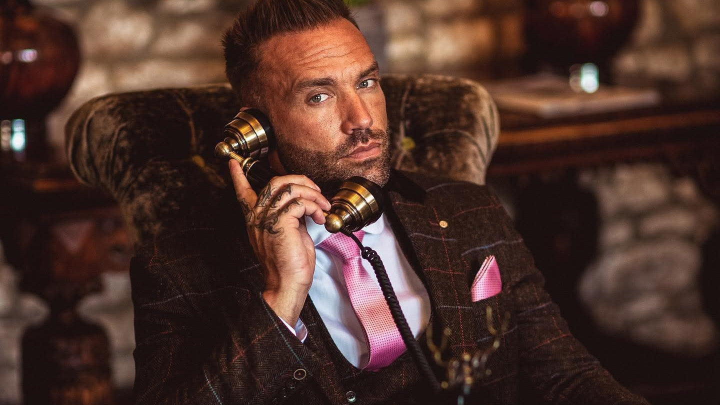 Callum Best in a tweed suit sitting in a chair, holding a vintage phone in a room with decorative elements.