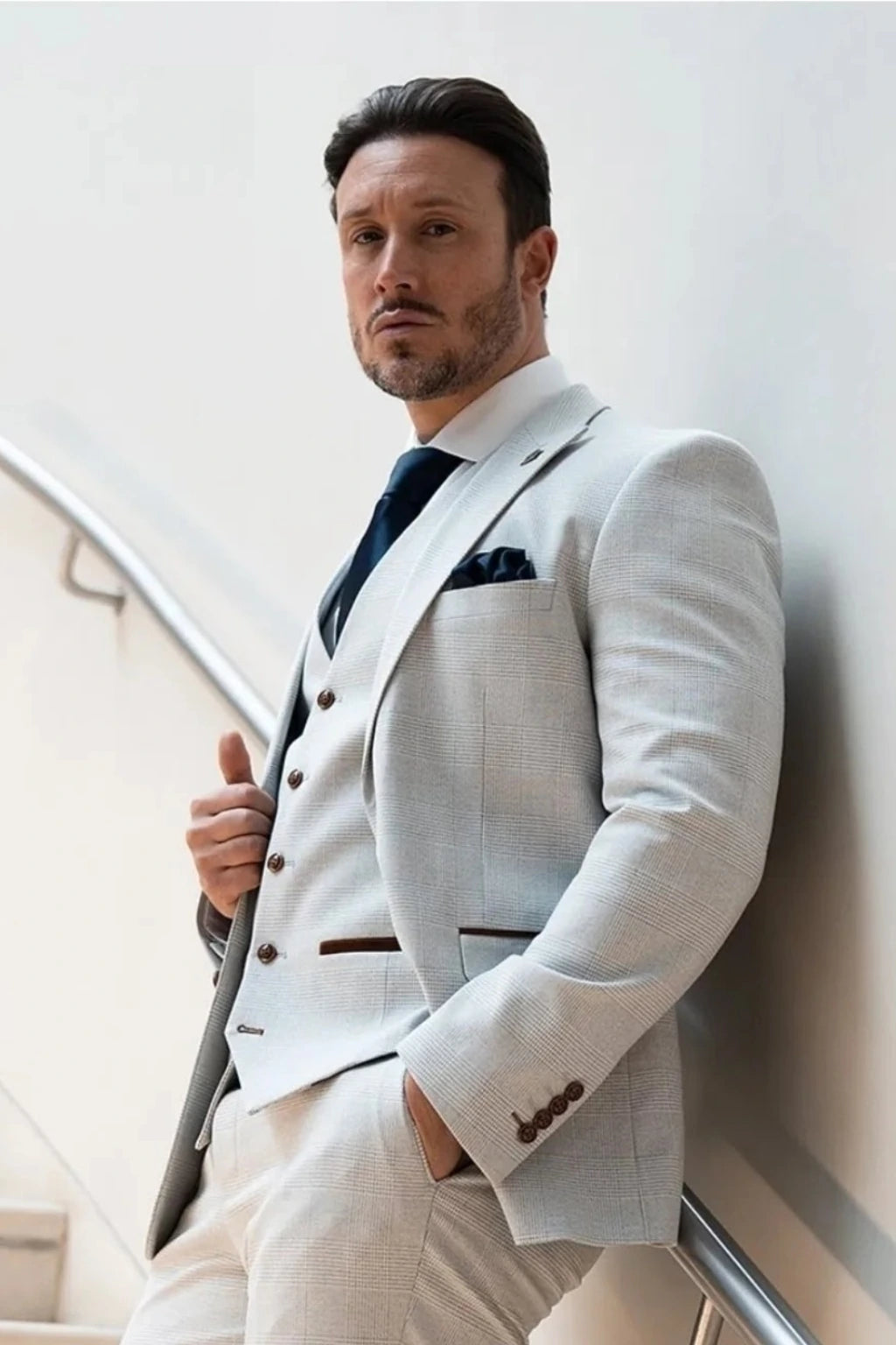 A man wearing the Paul Andrew Mark Stone three-piece suit with a matching vest and trousers, a white shirt, and a dark blue tie, leaning against a white wall on an indoor staircase.