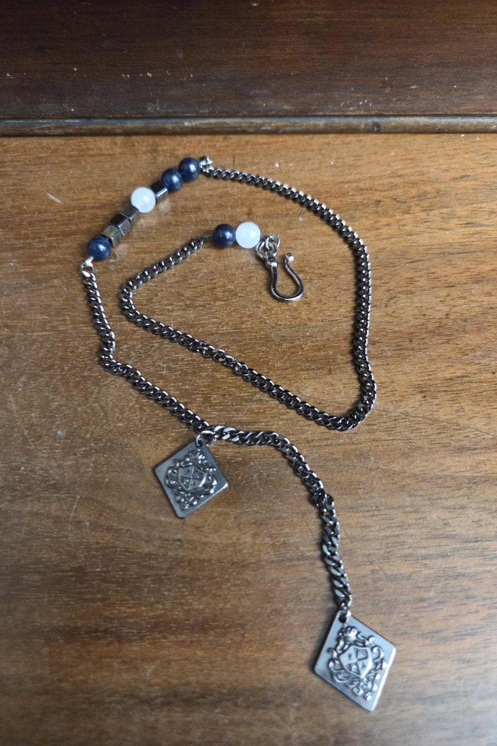 A silver-toned waistcoat with two square engraved pendants and a small beaded section with dark blue and white beads, displayed on a wooden surface.