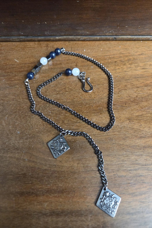 A silver-toned waistcoat with two square engraved pendants and a small beaded section with dark blue and white beads, displayed on a wooden surface.