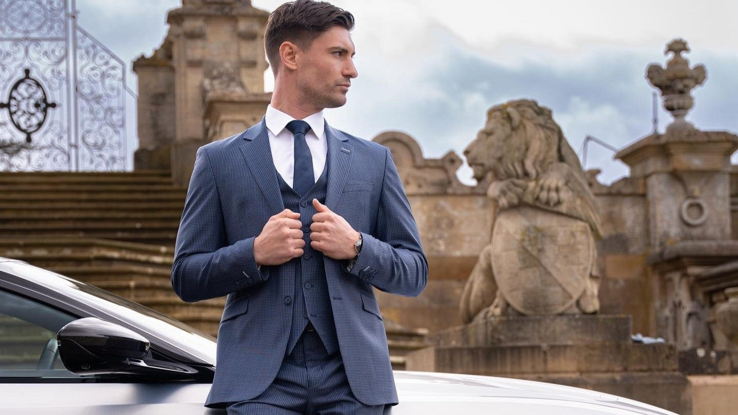 Man in a Cavani Bond Puppy Tooth 3-Piece Suit standing next to a Aston Martin car with a lion statue in the background.