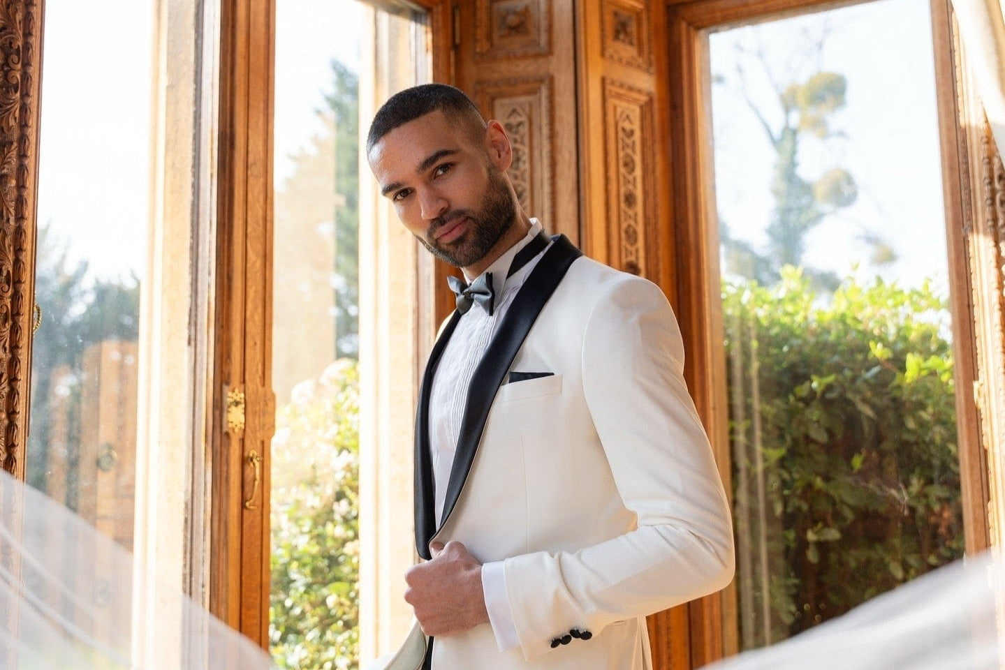 Man in a white tuxedo standing in a room with large windows and decorative walls.