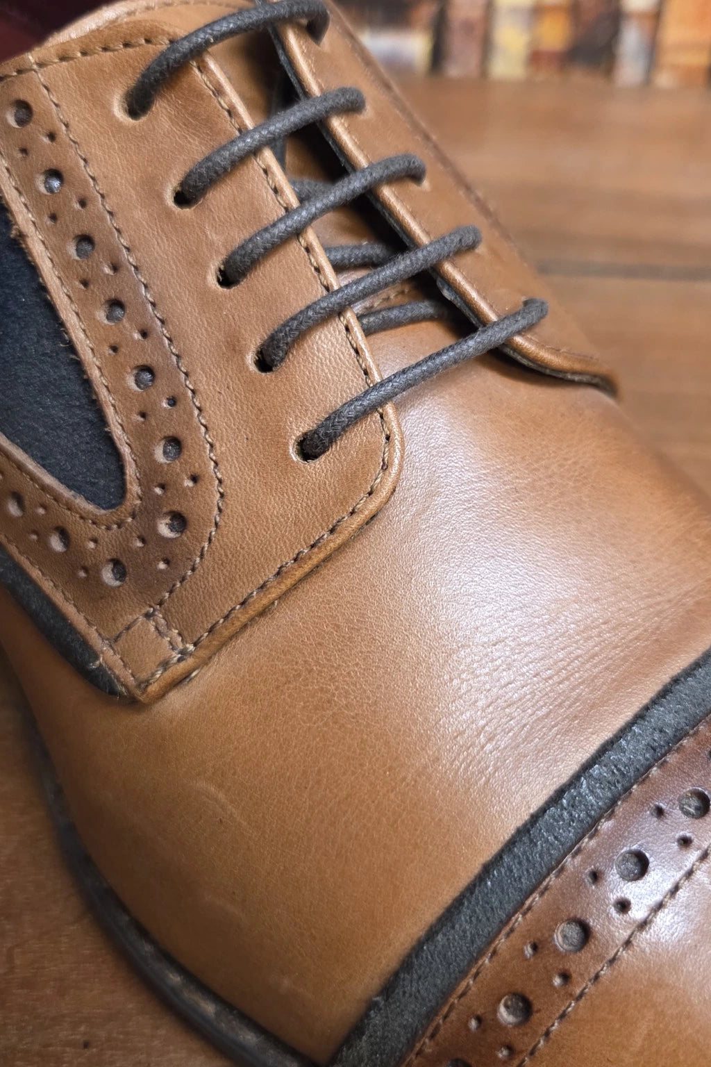 A close-up shot of a men's brown leather cap-toe oxford shoe with dark gray laces and brogue detailing, resting on a wooden surface.