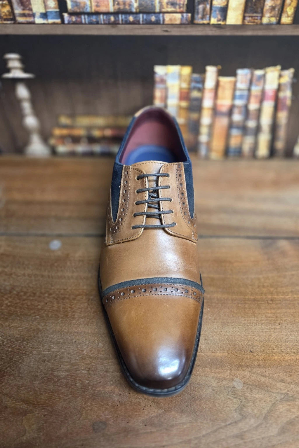 A men's brown leather cap-toe oxford shoe with dark navy blue side panels and laces, displayed on a wooden table with a blurred bookshelf background.
