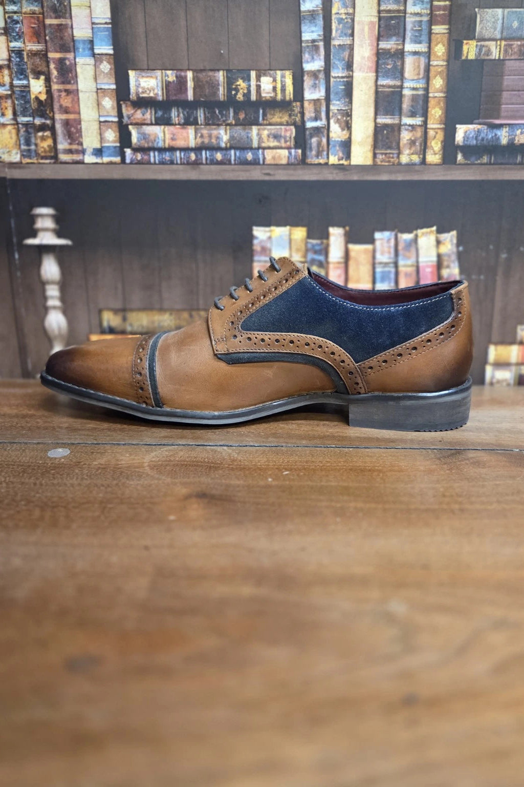 A men's brown leather cap-toe oxford shoe with dark blue suede wingtip and side panels, resting on a wooden table with a bookshelf in the background.