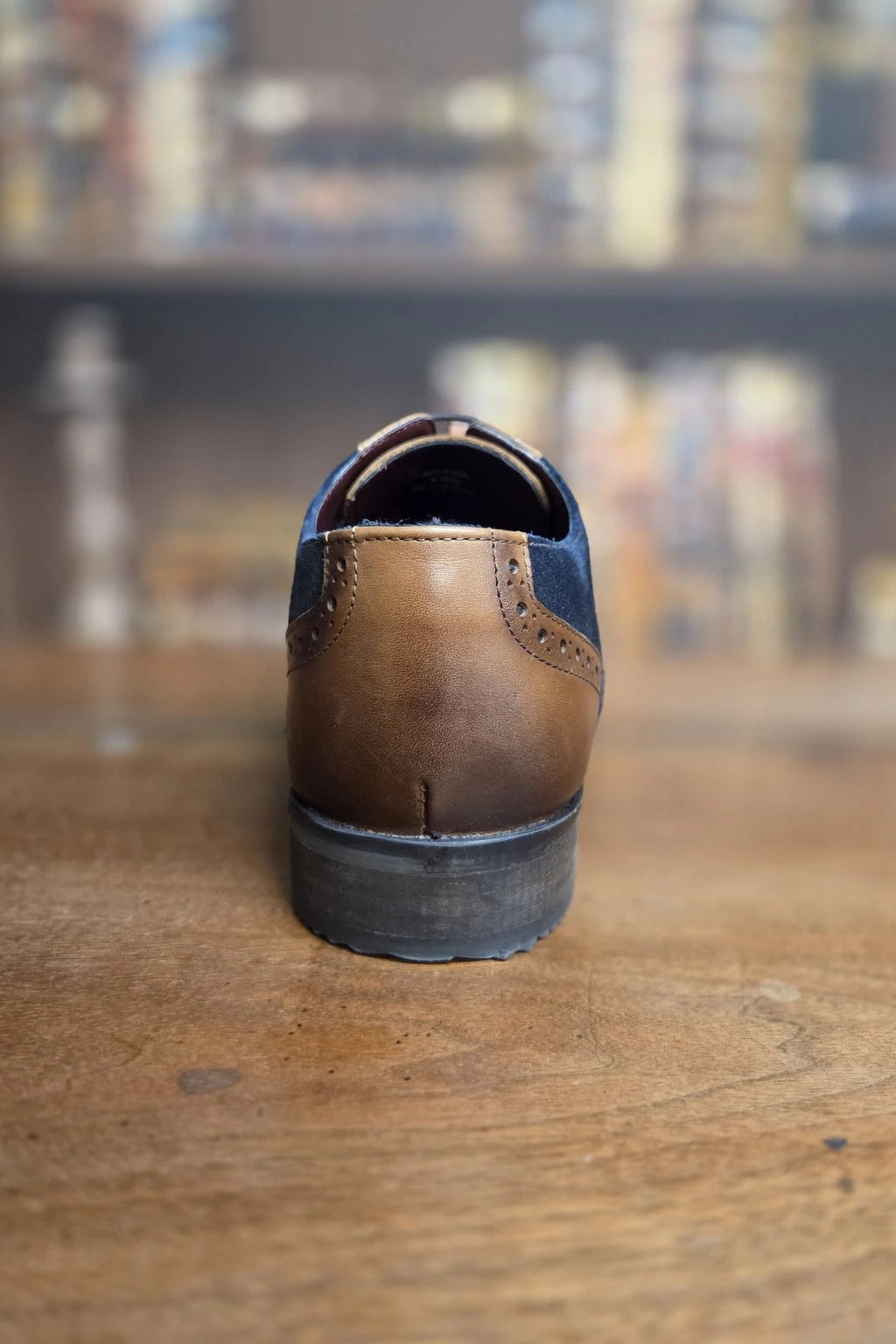 A brown leather wingtip shoe shown from the back, featuring dark blue suede side panels and a dark gray sole, positioned on a wooden table with a blurred bookshelf background.