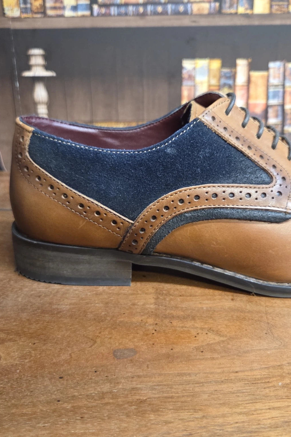 A brown and navy blue suede-and-leather wingtip oxford shoe resting on a wooden table, with a blurred background of bookshelves.