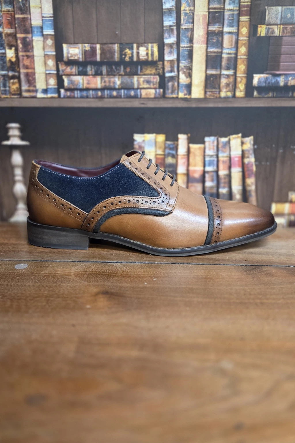 A men's cap-toe oxford shoe in tan leather and navy blue suede, positioned on a wooden table with a bookshelf in the background.