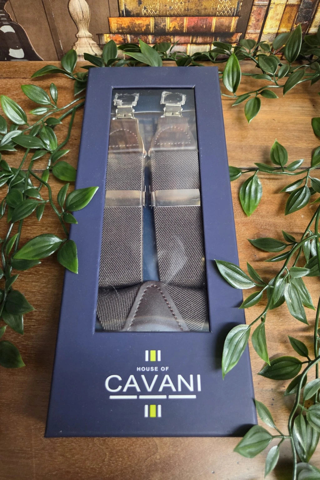 A pair of Cavani brown woven neck braces with clear metal loops, presented inside a navy blue Cavani gift box, styled on a wooden table with green leaves.