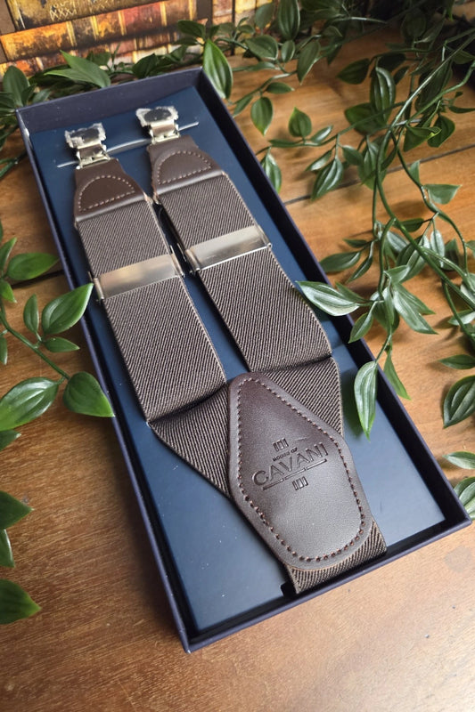 A pair of brown Cavani textured suspenders with silver metal clips, presented in a dark blue box on a wooden table with green leaves.