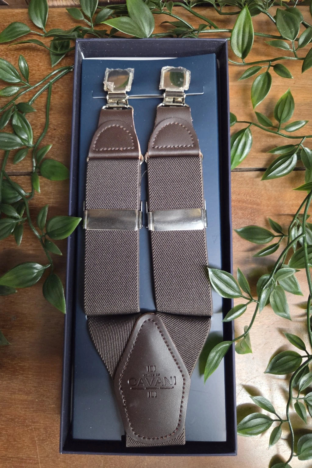 A pair of brown Cavani woven braces with silver metal clips, presented inside a dark blue gift box on a wooden surface surrounded by green leaves.