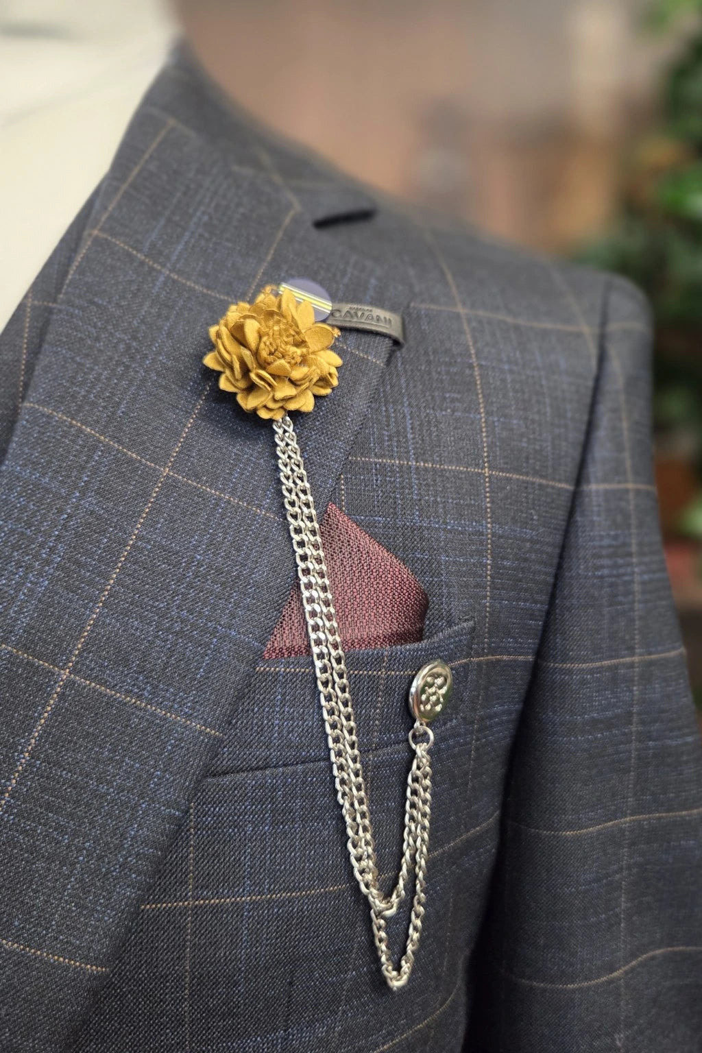 A close-up of a Cavani Torino navy windowpane check suit jacket featuring a gold flower lapel pin with a silver chain, a  and a Paul Andrew burgundy pocket square.