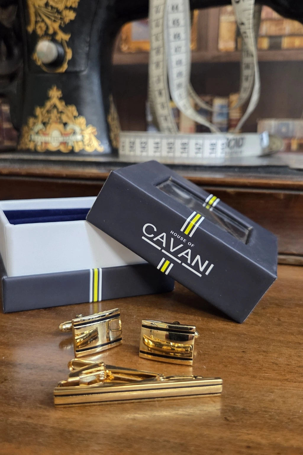 Gold-toned cufflinks and matching tie bar set displayed on a wooden table next to an open dark navy Cavani gift box, with a sewing machine and measuring tape blurred in the background.