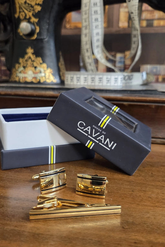 Gold-toned cufflinks and matching tie bar set displayed on a wooden table next to an open dark navy Cavani gift box, with a sewing machine and measuring tape blurred in the background.