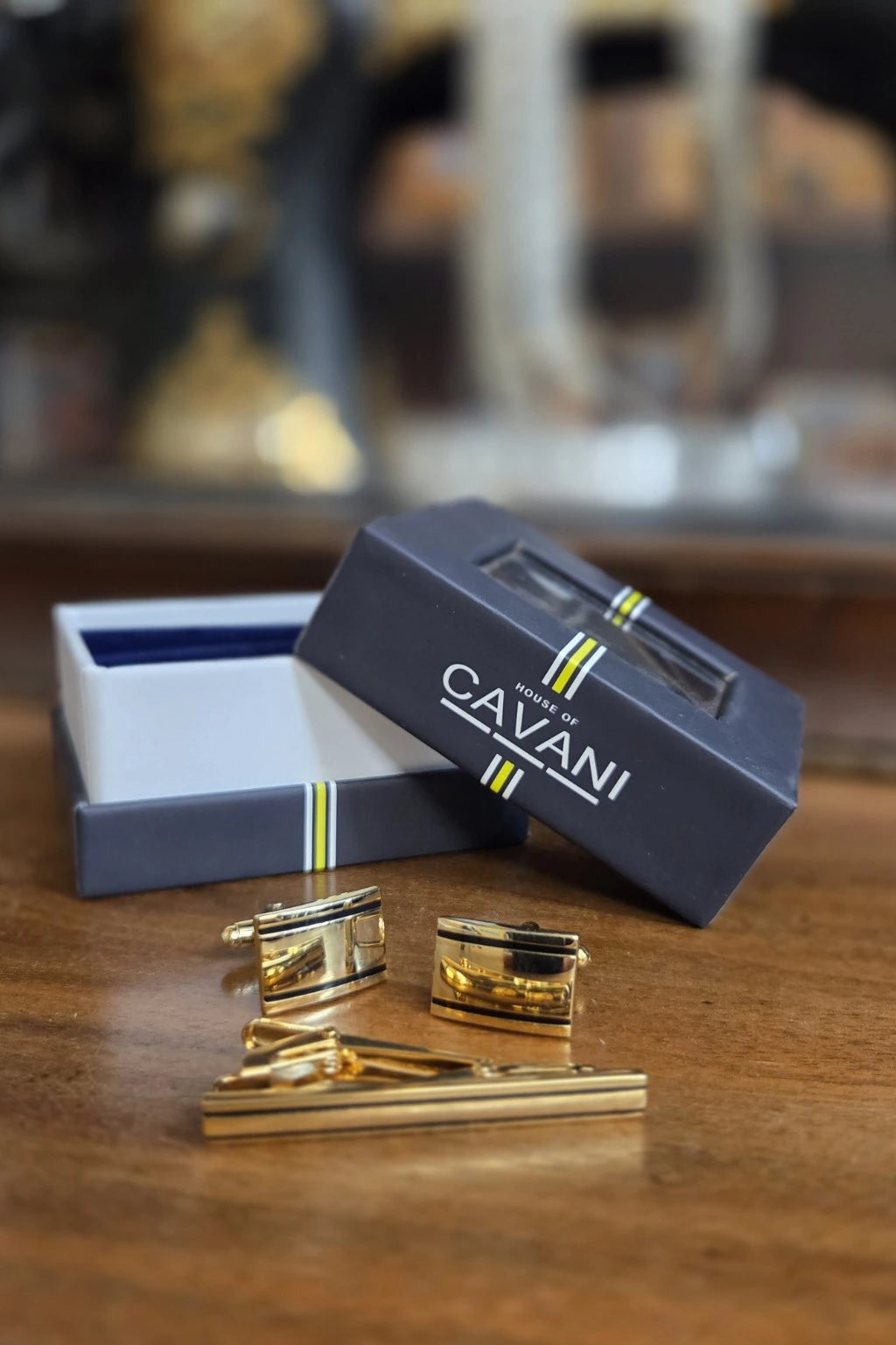A gold men's accessory set including cufflinks and a tie clip displayed in front of an open navy blue Cavani gift box on a wooden table.