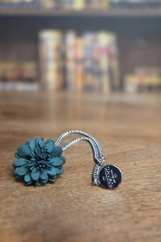 A silver-toned chain necklace featuring a textured dark teal flower charm and a small round black disc charm with white script, resting on a wooden tabletop with a blurred background.