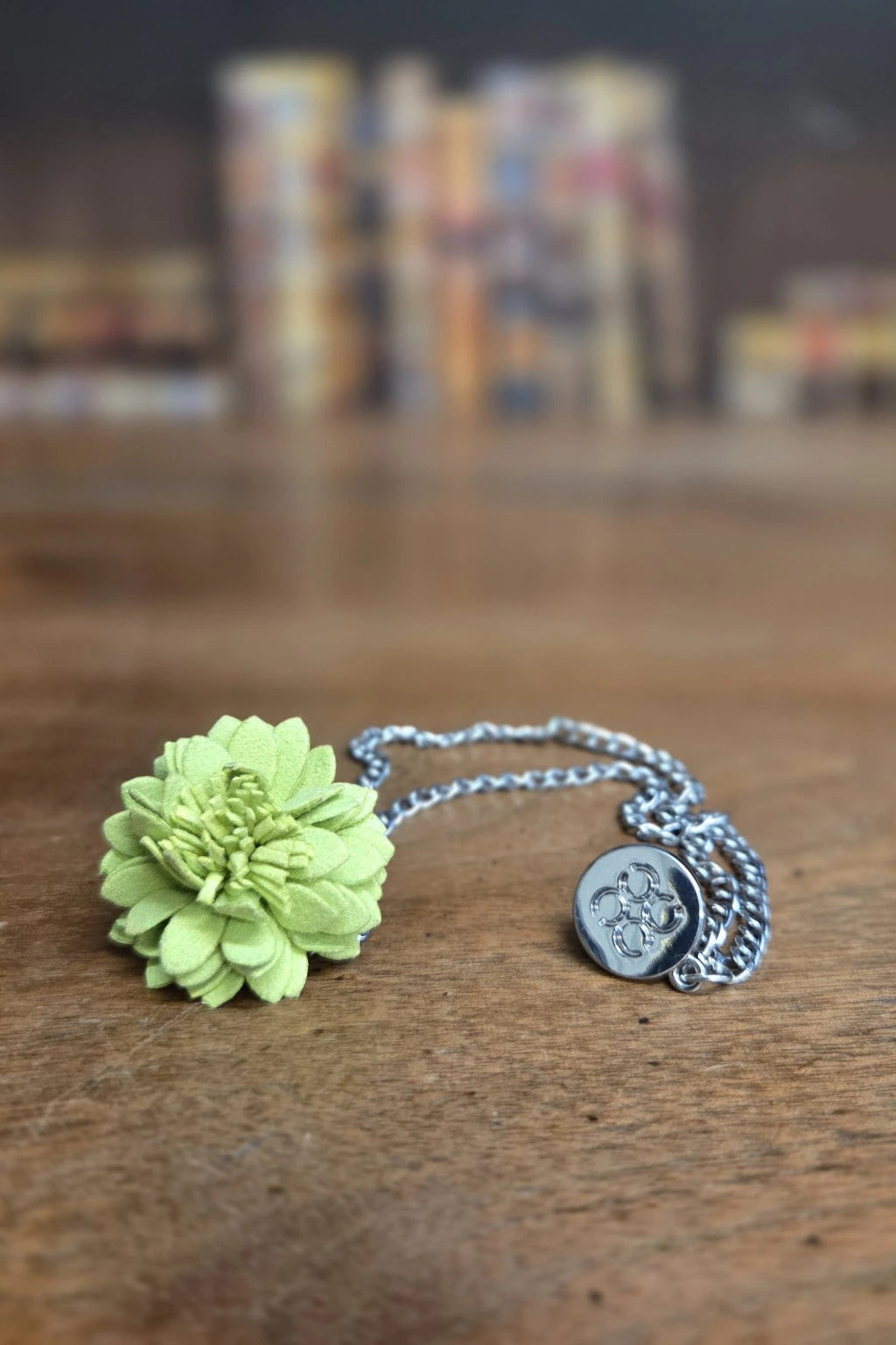 A lime green flower lapel pin, silver chain and a round silver Cavani medallion pendant, resting on a wooden tabletop with a blurred background.