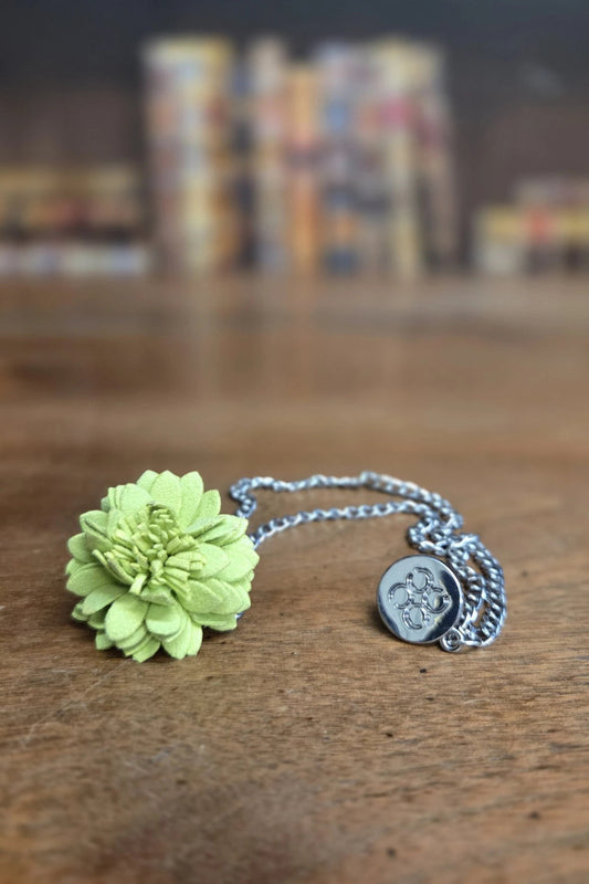 A silver chain necklace featuring a light green fabric flower charm and a small round silver coin pendant, displayed on a wooden surface with a blurred background.