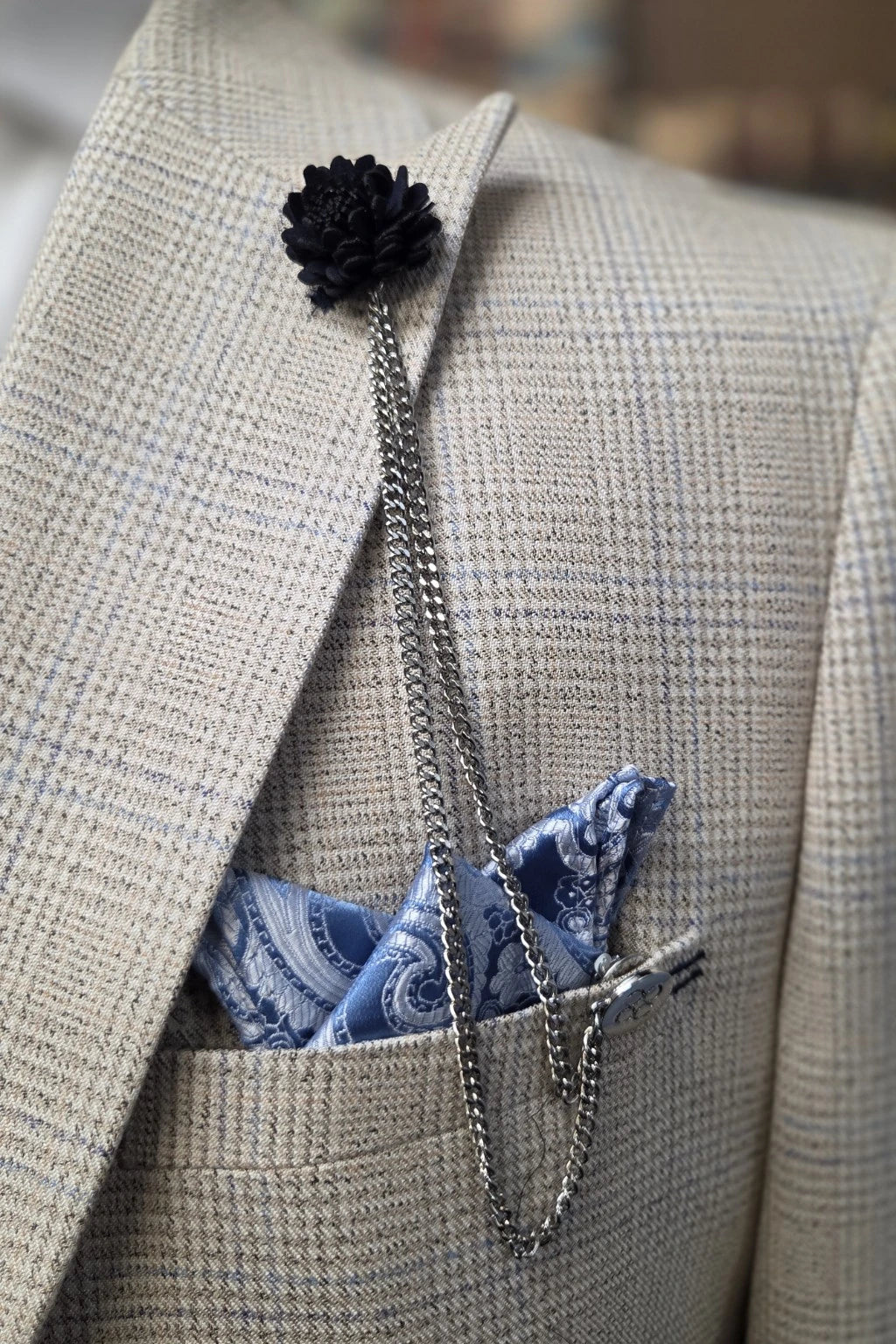 A close-up of a beige Caridi suit jacket with a navy flower lapel pin and a blue paisley pocket square with a silver chain tie.