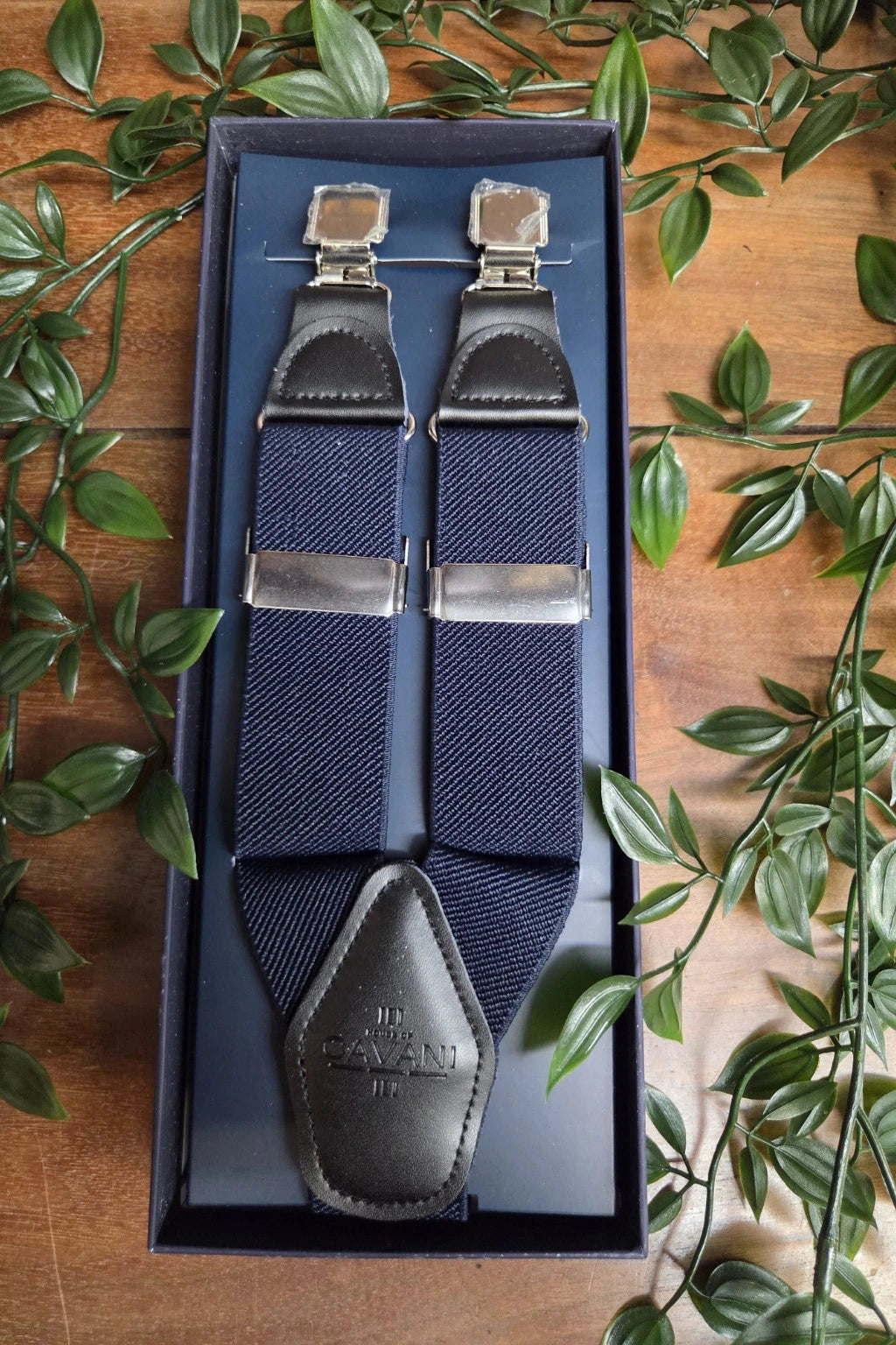 A pair of Cavani navy blue suspender straps with dark gray leather trim and silver metal clips, presented in a dark blue gift box on a wooden surface surrounded by green leaves.