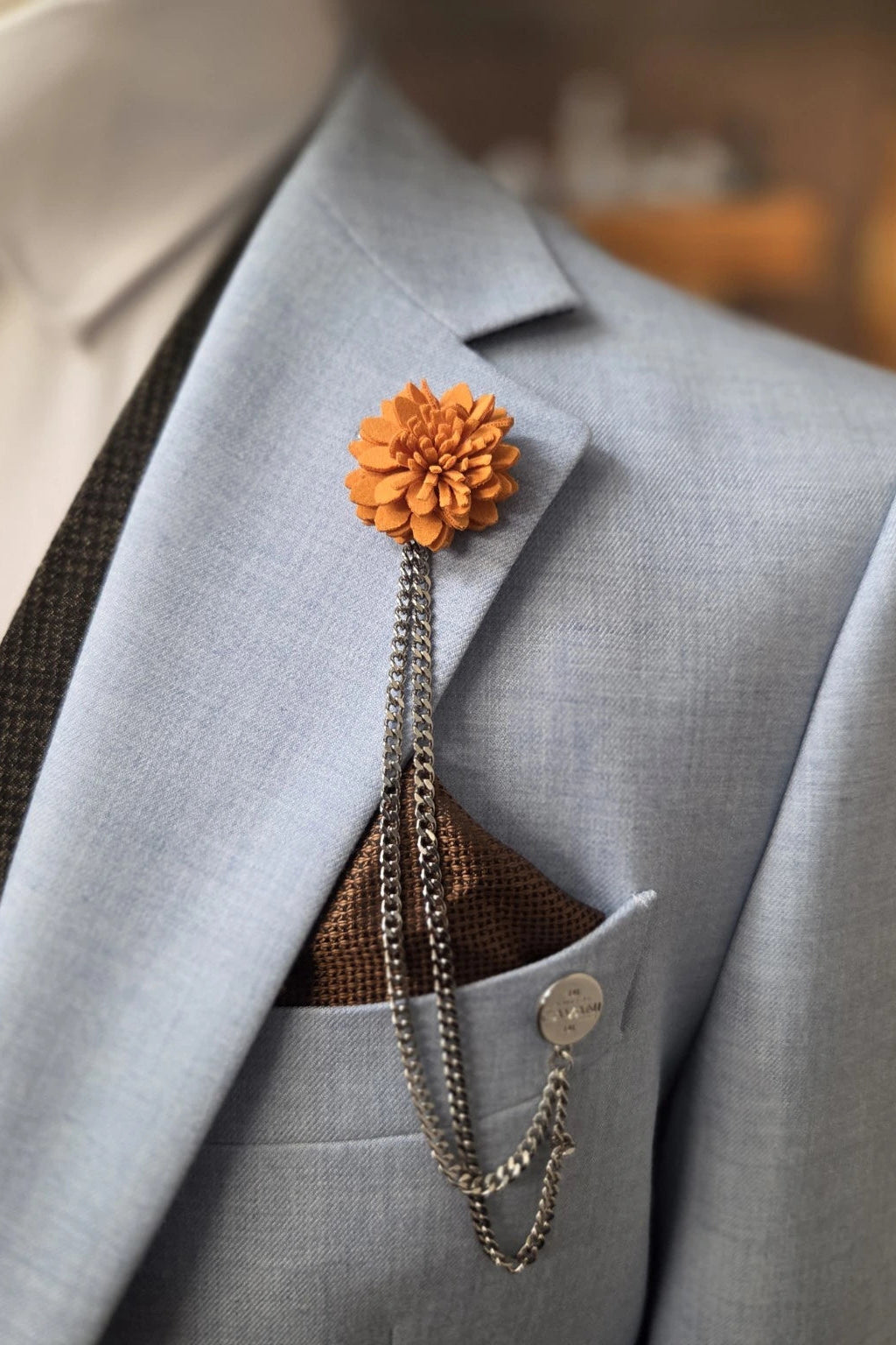 An orange flower lapel pin with a silver chain accessory, pinned to a light blue Malibu suit jacket over a Paul Andrew brown patterned pocket square.